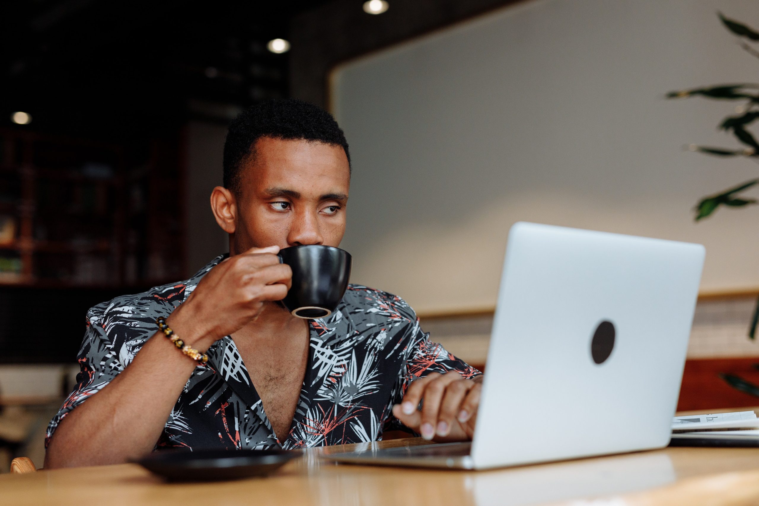 A man sits at a wooden desk and works on his laptop while sipping coffee from a mug.